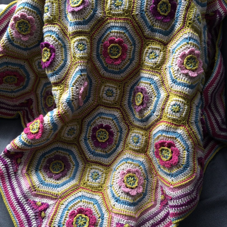 May include: A crocheted blanket with a hexagonal pattern featuring colorful flowers in shades of pink, purple, and green. The blanket is made with a variety of stitches and textures, creating a unique and eye-catching design.