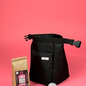 Op de afbeelding: Een zwarte canvas tas met een ritsvak en een verstelbare band. De tas ligt op een roze oppervlak naast een bruine papieren zak, een houten borstel en een klein rond blik met een roze etiket.