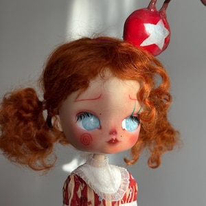 Può includere: Bambola fatta a mano con capelli rossi raccolti in due codini. La bambola ha grandi occhi blu, guance rosee e un vestito a fantasia rosso e bianco. Una palla rossa con una stella bianca è attaccata alla testa della bambola.