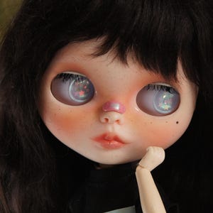 Può includere: Primo piano del viso di una bambola con grandi occhi riflettenti. La bambola ha i capelli scuri, le guance rosee e un piccolo naso rosa. Gli occhi riflettono un motivo colorato e stellato. La mano della bambola è vicino al mento.