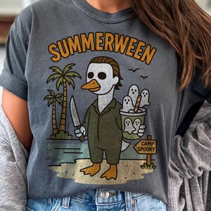 May include: Dark gray t-shirt with a graphic featuring a duck dressed as a horror movie character, holding a knife and a bucket of ghosts. The shirt reads "Summerween" in orange lettering, with a "Camp Spooky" sign.