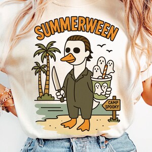 May include: Cream-colored t-shirt with the word "SUMMERWEEN" in orange lettering. The graphic features a duck dressed as a horror movie character holding a knife, with ghosts in a bucket and palm trees. The sign reads "CAMP SPOOKY".