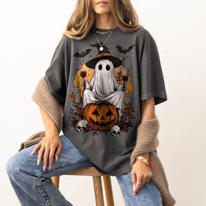 May include: Dark gray t-shirt featuring a Halloween-themed graphic. The design includes a ghost wearing a witch hat, holding wine glasses, and sitting on a carved pumpkin. Bats, candles, and skulls complete the spooky illustration.