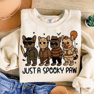 May include: Cream-colored sweatshirt featuring a graphic of four cartoon dogs dressed as horror movie characters. The phrase "JUST A SPOOKY PAW" is printed below the dogs. The dogs are wearing costumes of famous horror characters.