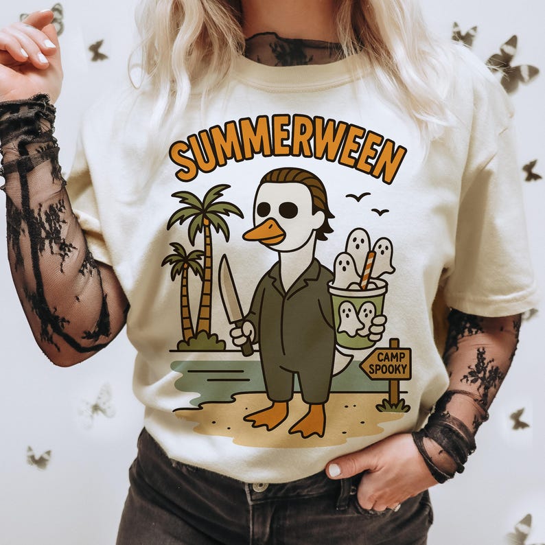 May include: Cream-colored t-shirt with the word "SUMMERWEEN" in orange lettering. The graphic features a duck wearing a Michael Myers mask and holding a knife, with ghosts in a bucket and palm trees.