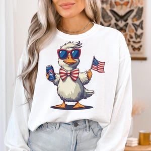 May include: White long-sleeved shirt with a cartoon duck wearing sunglasses, a bow tie, and holding a can and an American flag.