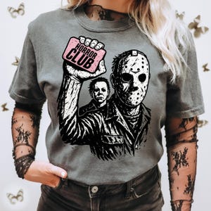 Pu&ograve; includere: T-shirt grigia con grafica di Jason Voorhees di Venerd&igrave; 13, che tiene in mano un cartello rosa con la scritta "HORROR CLUB". Il design include anche Michael Myers. Girocollo.