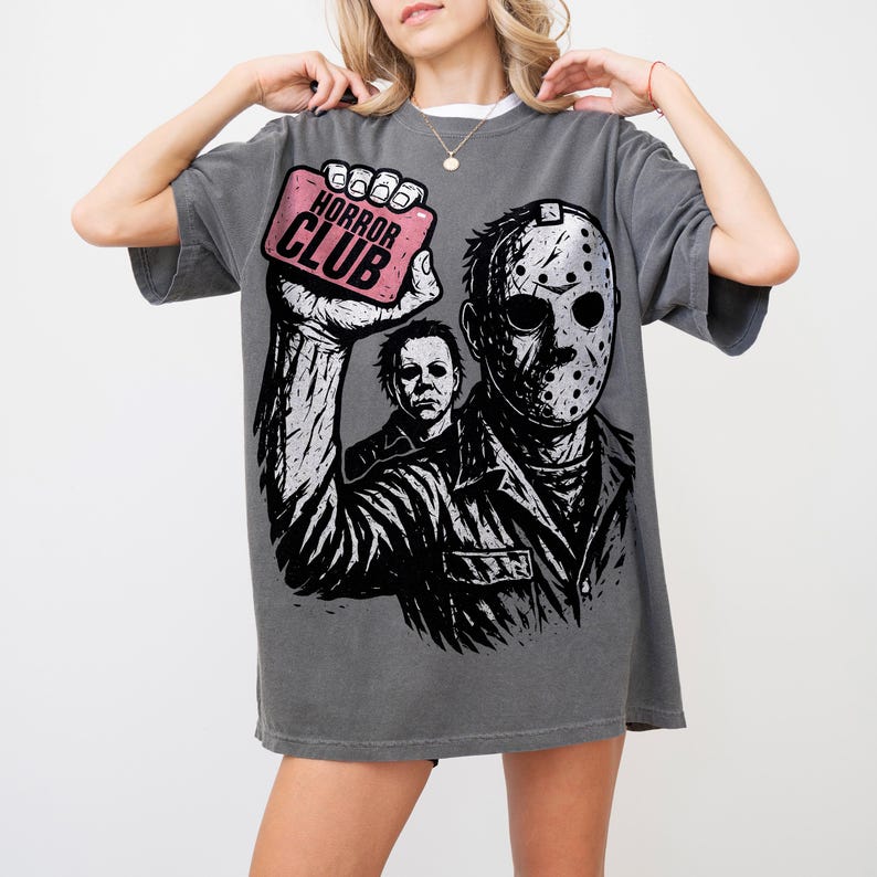 Pu&ograve; includere: T-shirt grigia con grafica di Jason Voorhees e Michael Myers. Il design include il testo "HORROR CLUB" su un rettangolo rosa. La maglietta ha una vestibilit&agrave; ampia.