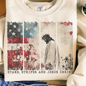 May include: Cream-colored sweatshirt featuring a distressed American flag design with red, white, and blue colors. The graphic includes an illustration of Jesus Christ and the text "STARS, STRIPES AND JESUS CHRIST".