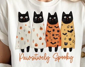 May include: White t-shirt featuring four black cats in Halloween costumes. The cats wear orange, white, and yellow outfits with pumpkin, bat, and ghost designs. The text "Pawsitively Spooky" is written in orange script below the cats.