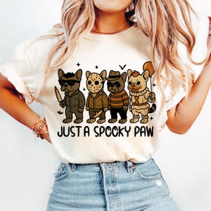May include: Cream-colored t-shirt featuring a graphic of four cartoon dogs dressed as horror movie characters. The dogs are in costumes, including a killer with a knife, and a clown holding a balloon. The text "JUST A SPOOKY PAW" is below the dogs.