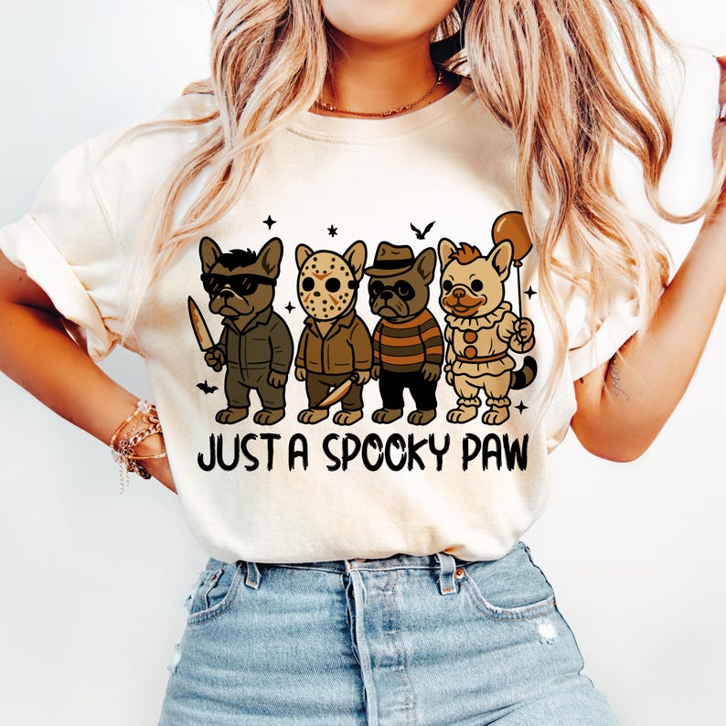May include: Cream-colored t-shirt featuring a graphic of four cartoon dogs dressed as horror movie characters. The dogs are in costumes, including a killer with a knife, and a clown holding a balloon. The text "JUST A SPOOKY PAW" is below the dogs.