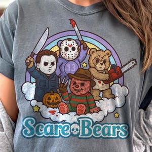 May include: A blue-grey t-shirt with a cartoon design of four teddy bears dressed as horror movie characters, including Michael Myers, Jason Voorhees, Freddy Krueger, and a bear with a chainsaw. The design includes a rainbow and the text "Scare Bears".