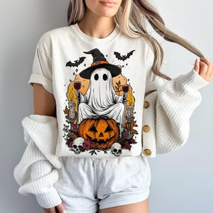 May include: Cream-colored t-shirt featuring a Halloween-themed graphic. The design includes a ghost wearing a witch hat, holding wine glasses, and sitting on a carved pumpkin. Bats, candles, and skulls complete the spooky illustration.
