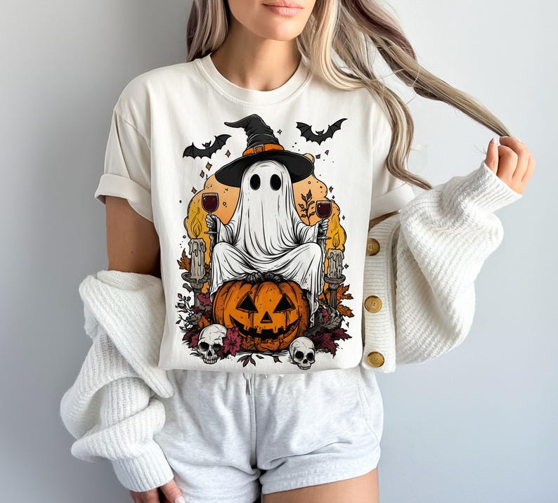 May include: Cream-colored t-shirt featuring a Halloween-themed graphic. The design includes a ghost wearing a witch hat, holding wine glasses, and sitting on a carved pumpkin. Bats, candles, and skulls complete the spooky illustration.