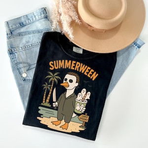 May include: Black t-shirt with the word "SUMMERWEEN" in orange lettering. The graphic features a duck-like character in a Michael Myers costume holding a knife and a bucket of ghosts. Light blue jeans and a tan hat are also in the image.