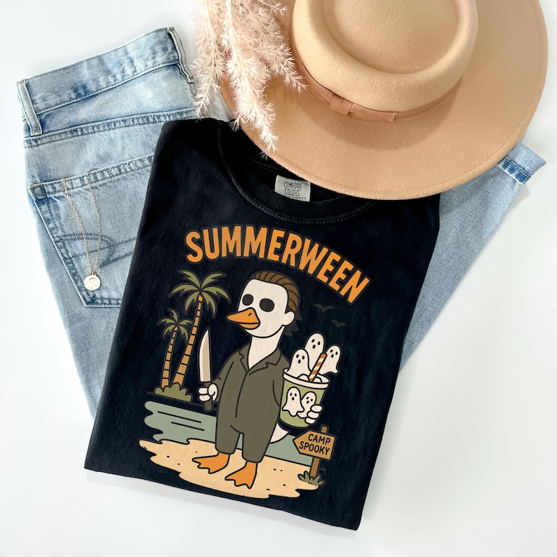May include: Black t-shirt with the word "SUMMERWEEN" in orange lettering. The graphic features a duck-like character in a Michael Myers costume holding a knife and a bucket of ghosts. Light blue jeans and a tan hat are also in the image.