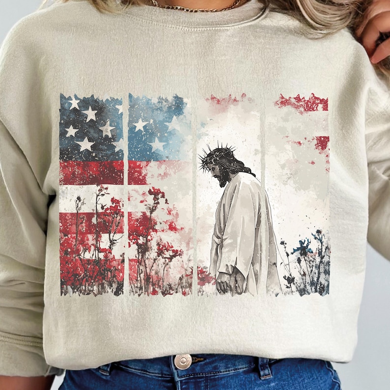 May include: A beige sweatshirt featuring a graphic of Jesus with a crown of thorns, set against a backdrop of the American flag and floral elements. The design incorporates red, white, and blue colors.