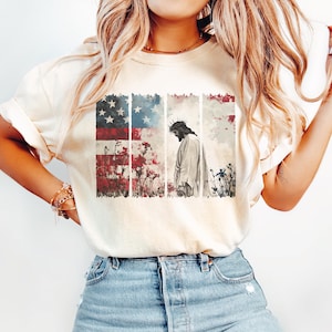 May include: Cream-colored t-shirt featuring a graphic design. The design includes an American flag, floral elements, and a depiction of Jesus wearing a crown of thorns. The shirt is paired with blue denim jeans.
