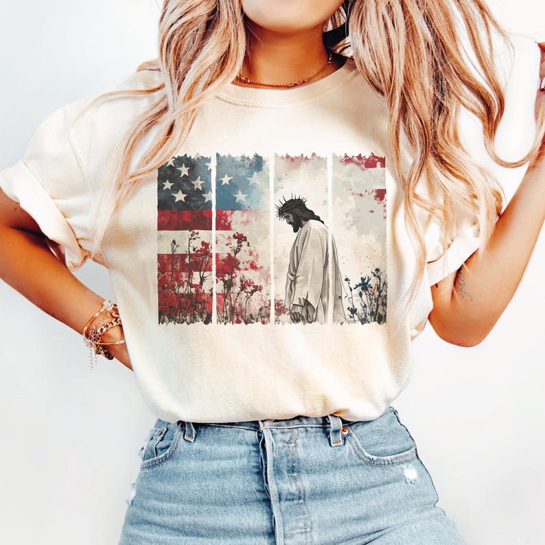 May include: Cream-colored t-shirt featuring a graphic design. The design includes an American flag, floral elements, and a depiction of Jesus wearing a crown of thorns. The shirt is paired with blue denim jeans.
