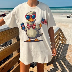 May include: White t-shirt with a cartoon duck wearing sunglasses and a red, white, and blue bow tie. The duck is holding a can of beer and a small American flag.
