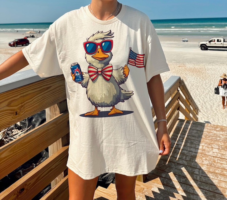 May include: White t-shirt with a cartoon duck wearing sunglasses and a red, white, and blue bow tie. The duck is holding a can of beer and a small American flag.