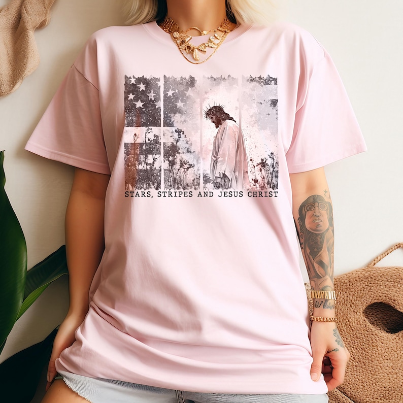 May include: A light pink t-shirt featuring a graphic design. The design includes an American flag motif with stars and stripes, a depiction of Jesus Christ, and the text "STARS, STRIPES AND JESUS CHRIST".