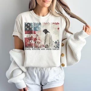 May include: Cream-colored t-shirt featuring a graphic of the American flag with a depiction of Jesus Christ. The text "STARS, STRIPES AND JESUS CHRIST" is printed below the image. The shirt is paired with gray shorts and a white cardigan.