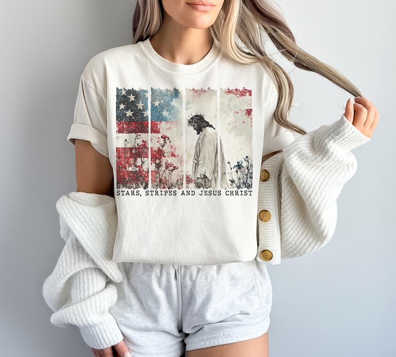 May include: Cream-colored t-shirt featuring a graphic of the American flag with a depiction of Jesus Christ. The text "STARS, STRIPES AND JESUS CHRIST" is printed below the image. The shirt is paired with gray shorts and a white cardigan.