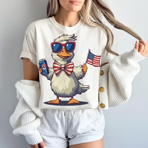 May include: White t-shirt with a cartoon duck wearing sunglasses and a red, white, and blue bow tie. The duck is holding a can of beer and an American flag.