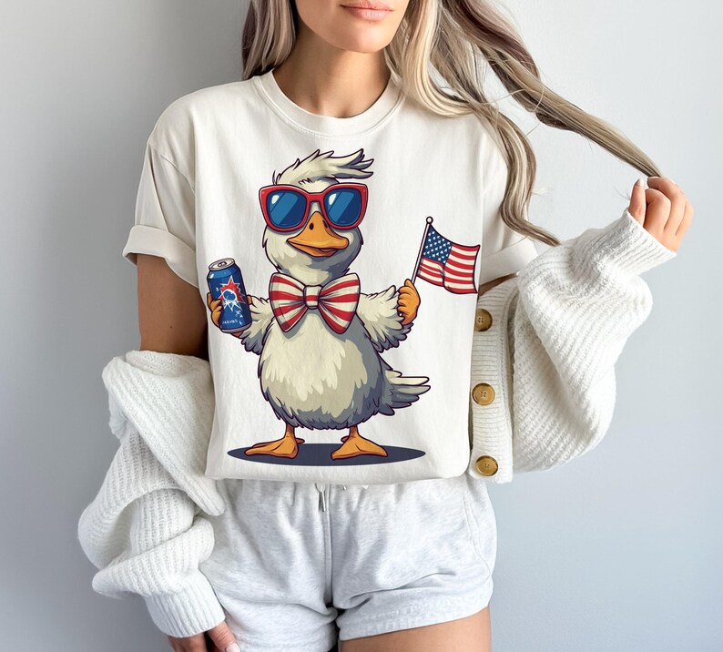 May include: White t-shirt with a cartoon duck wearing sunglasses and a red, white, and blue bow tie. The duck is holding a can of beer and an American flag.
