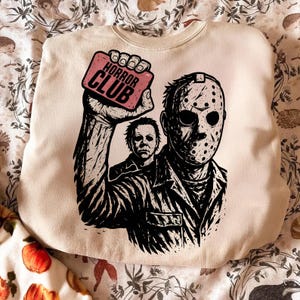 Pu&ograve; includere: Felpa color crema con grafica in bianco e nero di due personaggi di film horror. Un personaggio indossa una maschera da hockey, l'altro &egrave; sullo sfondo. La grafica include il testo "HORROR CLUB" su un rettangolo rosa.