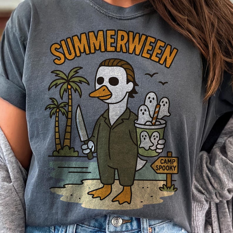 May include: Dark gray t-shirt with a humorous graphic. The design features a duck dressed as a horror movie character, holding a knife and a cup of ghost-shaped treats. The text "SUMMERWEEN" is arched above the image, with "CAMP SPOOKY" on a sign.