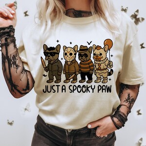 May include: Cream-colored t-shirt featuring a graphic of four cartoon dogs dressed as horror movie characters. The phrase "JUST A SPOOKY PAW" is printed below the dogs. The dogs are wearing costumes and holding props.