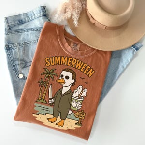May include: Rust-colored t-shirt with the word "SUMMERWEEN" in orange lettering. The graphic features a duck dressed as a horror movie character holding a knife and a bucket of ghosts. The shirt is paired with blue jeans and a tan hat.