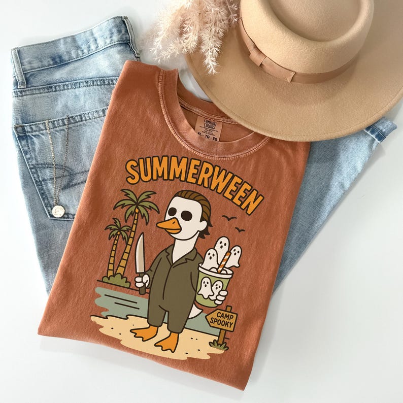 May include: Rust-colored t-shirt with the word "SUMMERWEEN" in orange lettering. The graphic features a duck dressed as a horror movie character holding a knife and a bucket of ghosts. The shirt is paired with blue jeans and a tan hat.
