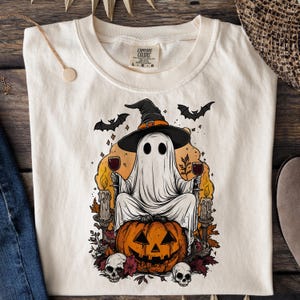 May include: Cream-colored t-shirt featuring a Halloween-themed graphic. The design includes a ghost wearing a witch hat, holding wine glasses, and sitting on a carved pumpkin. Bats, candles, and skulls complete the spooky scene.