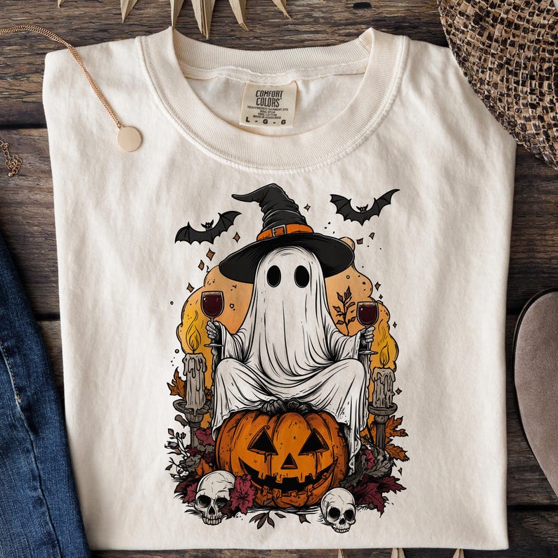 May include: Cream-colored t-shirt featuring a Halloween-themed graphic. The design includes a ghost wearing a witch hat, holding wine glasses, and sitting on a carved pumpkin. Bats, candles, and skulls complete the spooky scene.