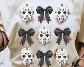 May include: Cream-colored t-shirt featuring a repeating pattern of hockey masks and black bows. The masks are white with black details, and the bows have lace-like accents. Red splatters are placed behind the masks and bows.