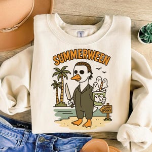 May include: Cream-colored sweatshirt with a graphic featuring a duck dressed as Michael Myers from Halloween, holding a knife and a cup of ghosts. The text "SUMMERWEEN" is arched above the illustration, with palm trees and a "Camp Spooky" sign.