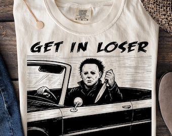 Puede incluir: Camiseta color crema con un gráfico de Michael Myers en un coche, sosteniendo un cuchillo. El texto dice "GET IN LOSER WE'RE GOING SLASHING". La camiseta tiene cuello redondo y mangas cortas. Se ve una etiqueta Comfort Colors.