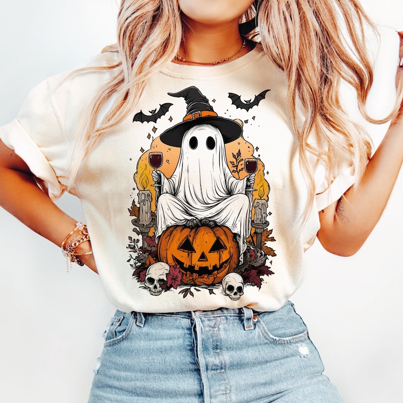 May include: Cream-colored t-shirt featuring a Halloween-themed graphic. The design includes a ghost wearing a witch hat, sitting on a carved pumpkin, holding wine glasses, with bats and skulls. The graphic is in shades of orange, black, and white.