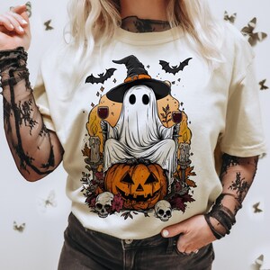 May include: Cream-colored t-shirt featuring a Halloween-themed graphic. The design includes a ghost wearing a witch hat, holding wine glasses, and sitting on a carved pumpkin. Bats, skulls, and candles complete the spooky illustration.