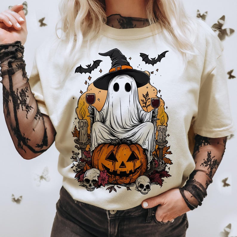 May include: Cream-colored t-shirt featuring a Halloween-themed graphic. The design includes a ghost wearing a witch hat, holding wine glasses, and sitting on a carved pumpkin. Bats, skulls, and candles complete the spooky illustration.