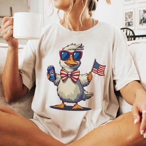 May include: White t-shirt with a cartoon duck wearing sunglasses and a red, white, and blue bow tie. The duck is holding a can of beer and a small American flag.