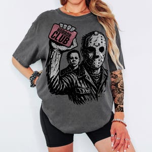 Pu&ograve; includere: T-shirt grigio scuro con una grafica di Jason Voorhees e Michael Myers. Il design include il testo "HORROR CLUB" su un rettangolo rosa. La maglietta ha un aspetto vintage e usurato. Un capo d'abbigliamento casual.