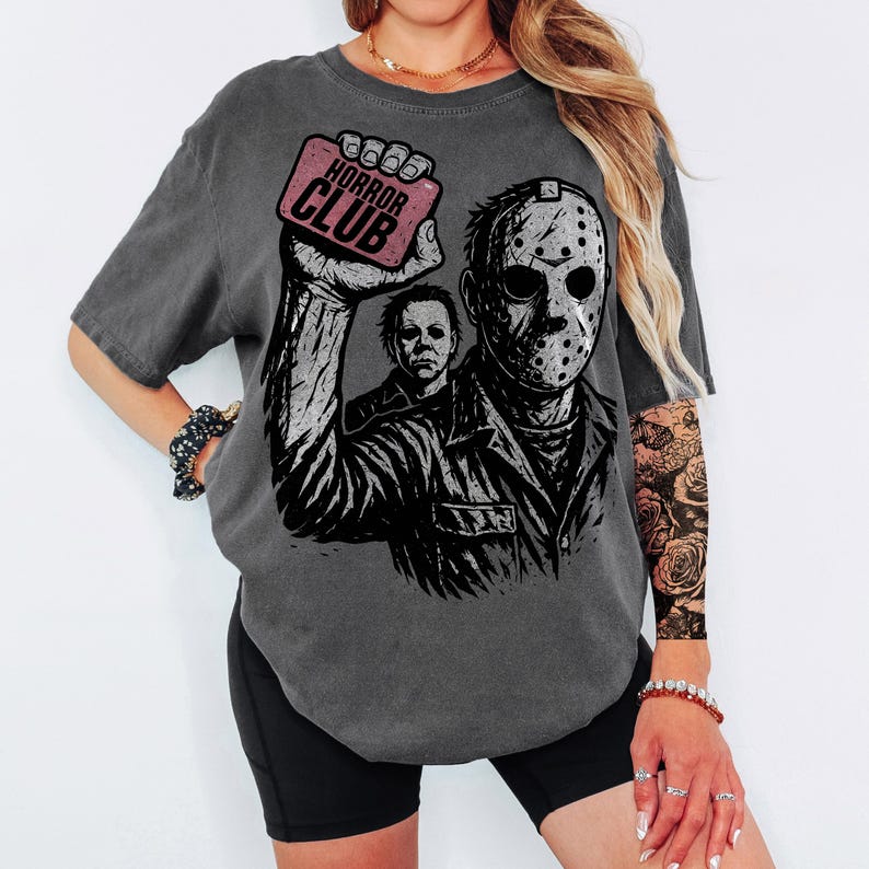 Pu&ograve; includere: T-shirt grigio scuro con una grafica di Jason Voorhees e Michael Myers. Il design include il testo "HORROR CLUB" su un rettangolo rosa. La maglietta ha un aspetto vintage e usurato. Un capo d'abbigliamento casual.