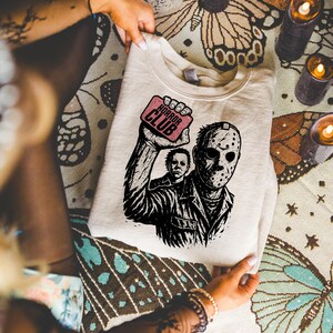 Pu&ograve; includere: Felpa color crema con una grafica in bianco e nero di personaggi di film horror, tra cui una figura mascherata e un altro personaggio. La grafica include il testo "HORROR CLUB" su un rettangolo rosa. La felpa &egrave; piegata.