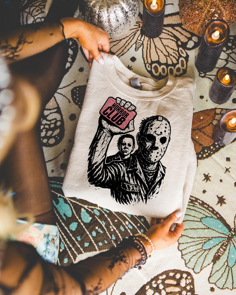 Pu&ograve; includere: Felpa color crema con una grafica in bianco e nero di personaggi di film horror, tra cui una figura mascherata e un altro personaggio. La grafica include il testo "HORROR CLUB" su un rettangolo rosa. La felpa &egrave; piegata.