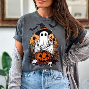May include: Dark gray t-shirt featuring a Halloween-themed graphic. The design includes a ghost wearing a witch hat, sitting on a carved pumpkin, holding wine glasses, with bats and skulls. The shirt is made of a soft material.